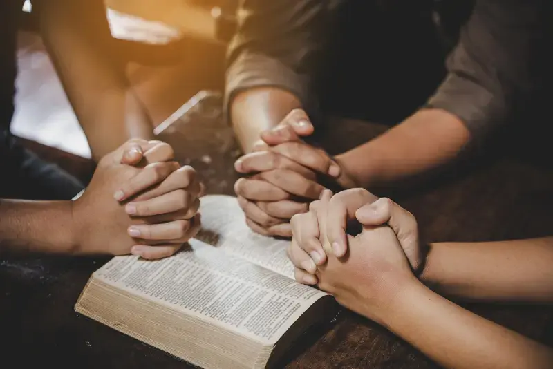 three sets of hands clasped in prayer on top of a bible
